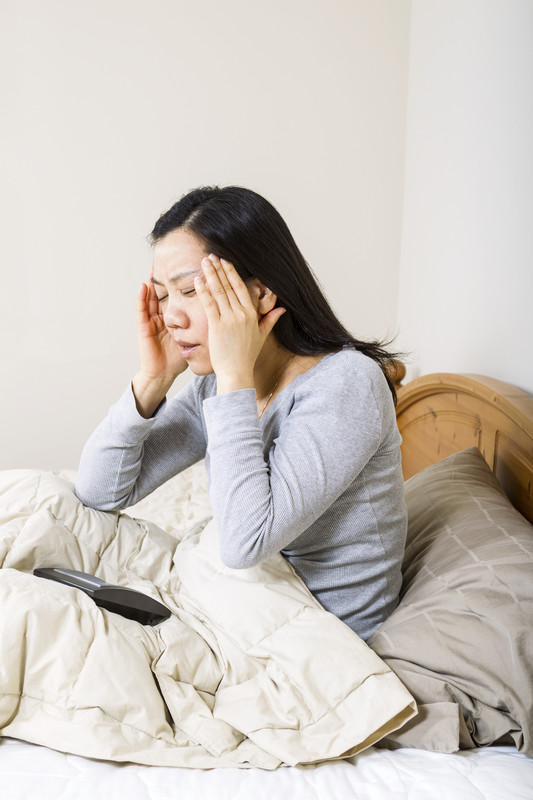 Image of a mature woman, holding her head with both hands, while alarm clock is lying in bed